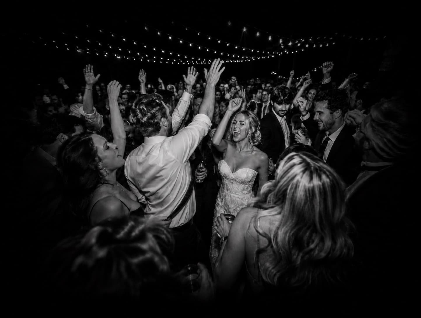 A packed wedding dance floor under string lights, hands in the air, shot in black and white